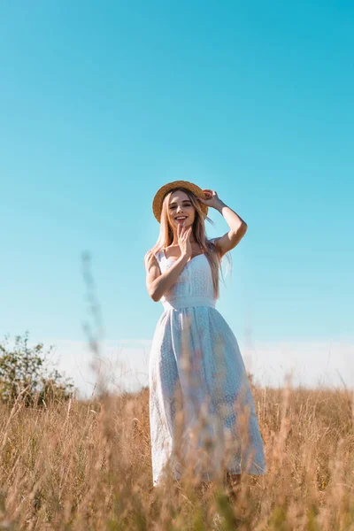 Selective focus of blonde woman in white dress touching straw hat and holding hand near face while standing in field — Stock Photo