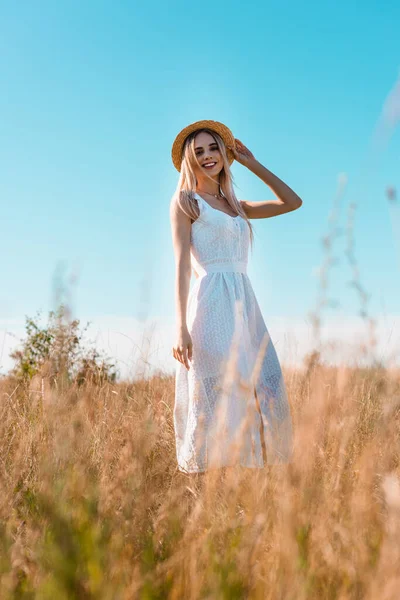 Foco seletivo da jovem mulher loira em vestido branco tocando chapéu de palha enquanto posando em campo contra o céu azul — Fotografia de Stock