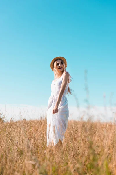 Selective focus of blonde woman in white dress and straw hat standing in field against blue sky and looking away — Stock Photo