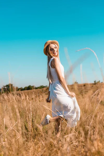 Selective focus of woman in white dress and straw hat standing on one leg while posing in meadow — Stock Photo