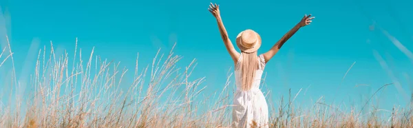 Selective focus of blonde woman in white dress and straw hat standing with raised hands against blue sky, horizontal image — Stock Photo