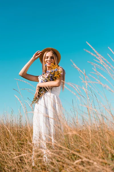 Selective focus of young blonde woman in white dress holding wildflowers and touching straw hat while looking at camera — Stock Photo