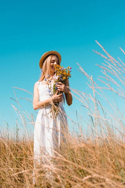 Fuoco selettivo della donna bionda in cappello di paglia e vestito bianco che tiene bouquet di fiori selvatici contro il cielo blu — Foto stock