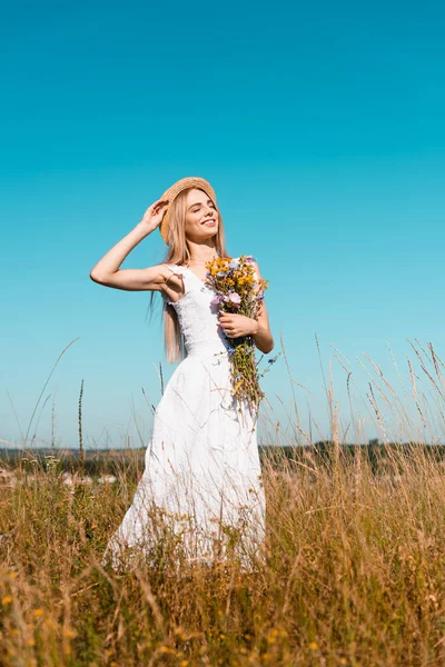 Selective focus of sensual woman in white dress touching straw hat and holding wildflowers in grassy field — Stock Photo