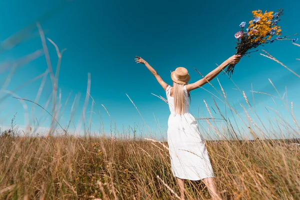 Back view of woman in white dress and straw hat holding wildflowers while standing with outstretched hands, selective focus — Stock Photo