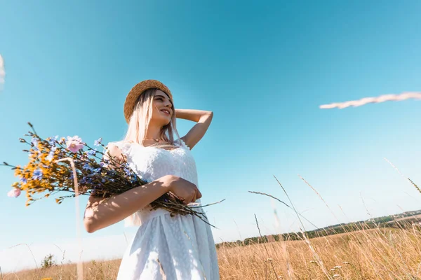 Vista ad alto angolo di giovane donna con fiori di campo toccando cappello di paglia mentre guardando lontano contro il cielo blu — Foto stock