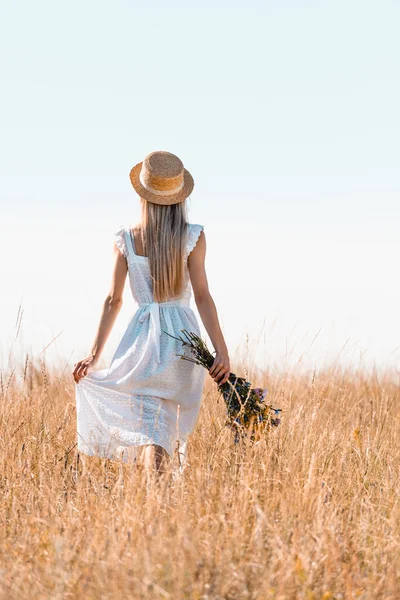 Vue arrière de la femme en chapeau de paille touchant robe blanche tout en marchant dans prairie herbeuse avec bouquet de fleurs sauvages — Photo de stock