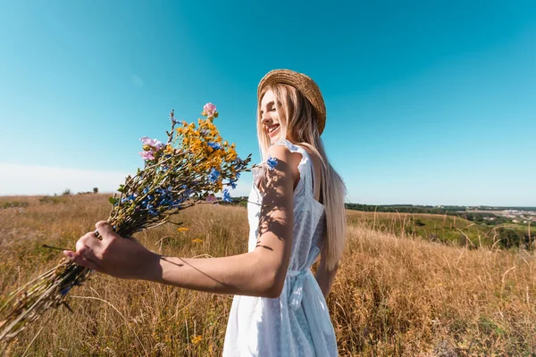 Femme blonde en chapeau de paille et robe blanche tenant bouquet de fleurs sauvages dans la prairie contre le ciel bleu — Photo de stock