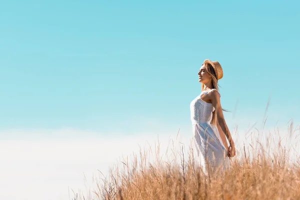 Side view of young woman in white dress and straw hat standing with closed eyes on grassy hill — Stock Photo