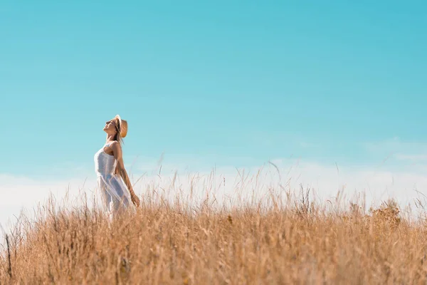 Selective focus of young woman in white dress and straw hat standing with outstretched hands and raised head against blue sky — Stock Photo