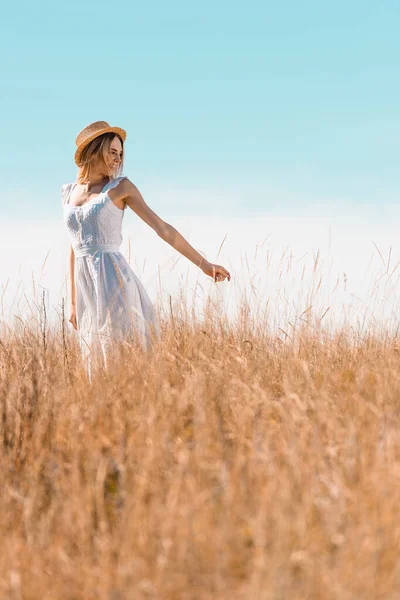 Selective focus of blonde woman in white dress and straw hat showing follow me gesture in grassy meadow — Stock Photo