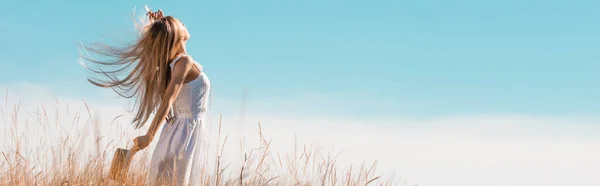 Horizontales Bild einer blonden Frau im weißen Kleid mit Strohhut, während sie mit erhobener Hand auf einem grasbewachsenen Hügel steht — Stockfoto
