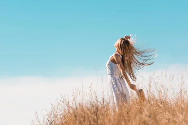 Foyer sélectif de la femme blonde en robe blanche tenant chapeau de paille tout en se tenant debout avec la main levée sur la colline herbeuse — Photo de stock