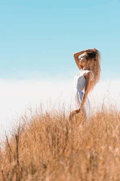 Селективное внимание молодой женщины в белом платье касаясь волос и держа соломенную шляпу, стоя на травянистом поле с закрытыми глазами — стоковое фото