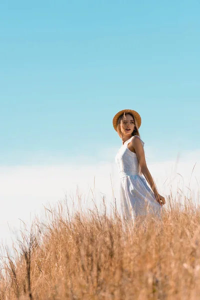 Foyer sélectif de la jeune femme en chapeau de paille touchant robe blanche tout en se tenant debout sur la colline herbeuse — Photo de stock