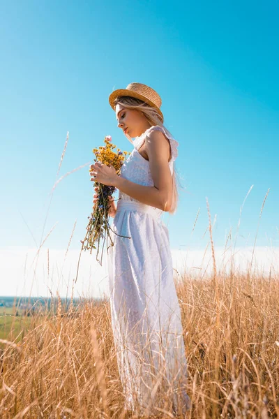 Side view of stylish woman in white dress and straw hat holding wildflowers against blue sky — Stock Photo