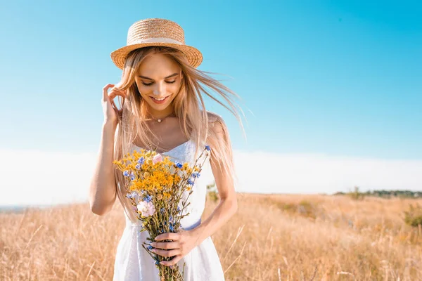 Sensual young woman in white dress and straw hat holding wildflowers and touching hair in grassy meadow — Stock Photo