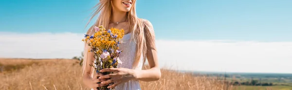 Vue recadrée de la femme blonde tenant des fleurs sauvages contre le ciel bleu, image horizontale — Photo de stock