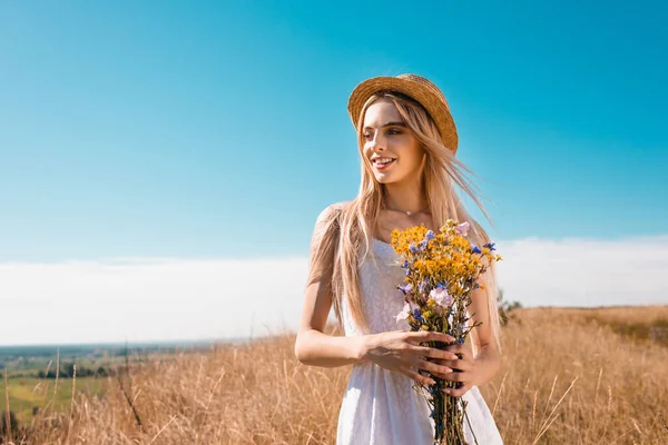 Blonde woman in white dress and straw hat holding bouquet of wildflowers while looking away against blue sky — Stock Photo