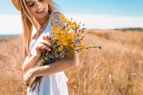 Selective focus of young blonde woman holding bouquet of wildflowers in meadow — Stock Photo