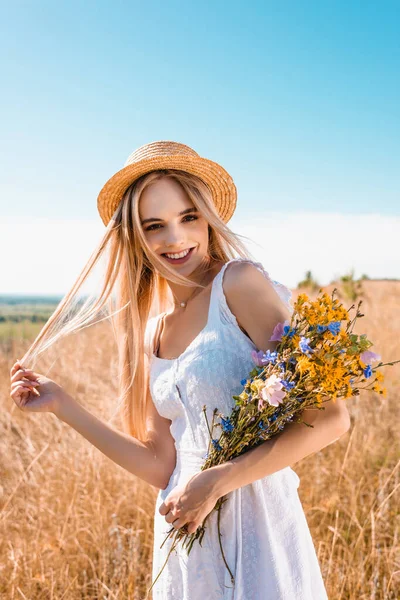 Jovem mulher elegante em chapéu de palha e vestido branco segurando flores silvestres e tocando o cabelo enquanto olha para a câmera — Fotografia de Stock