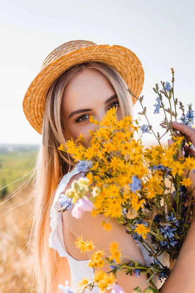 Чуттєва блондинка в солом'яному капелюсі тримає дикі квіти і дивиться на камеру проти неба — стокове фото