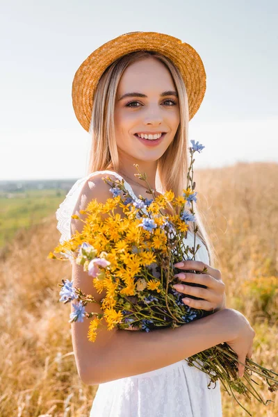 Junge sinnliche Frau in weißem Kleid und Strohhut blickt in die Kamera, während sie Wildblumen auf der Wiese hält — Stockfoto