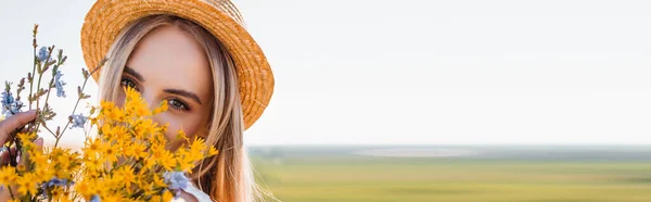 Plano panorámico de mujer rubia en sombrero de paja sosteniendo flores silvestres y mirando a la cámara contra el cielo despejado y el campo verde - foto de stock