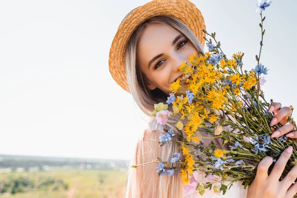 Mulher loira de chapéu de palha segurando flores silvestres enquanto olha para a câmera contra o céu claro — Fotografia de Stock