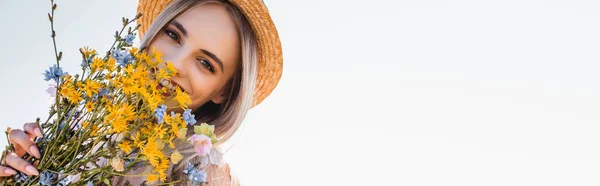 Concepto horizontal de mujer joven en sombrero de paja mirando a la cámara mientras sostiene flores silvestres contra el cielo despejado - foto de stock