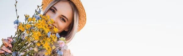 Conceito horizontal de jovem mulher em chapéu de palha olhando para a câmera enquanto segurando flores silvestres contra o céu claro — Fotografia de Stock