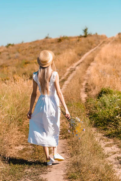 Vue arrière de la jeune femme en robe blanche et chapeau de paille tenant des fleurs sauvages tout en marchant sur la route dans la prairie — Photo de stock