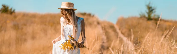 Horizontal image of blonde woman in straw hat touching white dress while standing in field with bouquet of wildflowers — Stock Photo