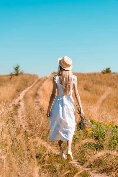 Vista trasera de mujer joven en vestido blanco y sombrero de paja caminando en el campo con ramo de flores silvestres - foto de stock