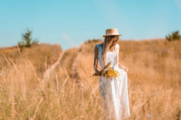 Giovane donna bionda in cappello di paglia toccare vestito bianco mentre in piedi in campo con bouquet di fiori di campo — Foto stock