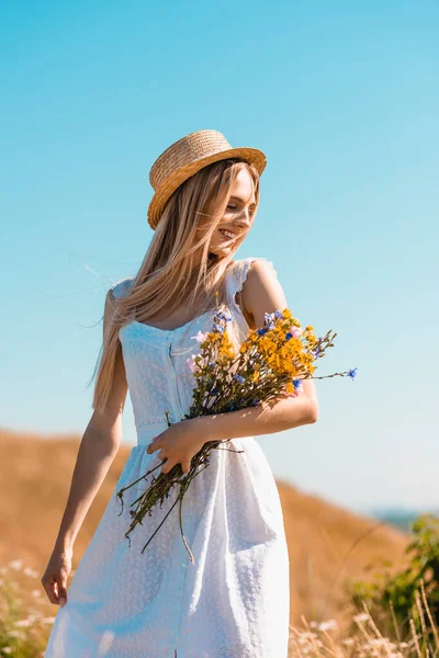 Mulher sensual em vestido branco e chapéu de palha segurando buquê de flores silvestres contra o céu azul — Fotografia de Stock