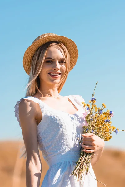 Jovem mulher em vestido branco e chapéu de palha segurando buquê de flores silvestres e olhando para a câmera contra o céu azul — Fotografia de Stock