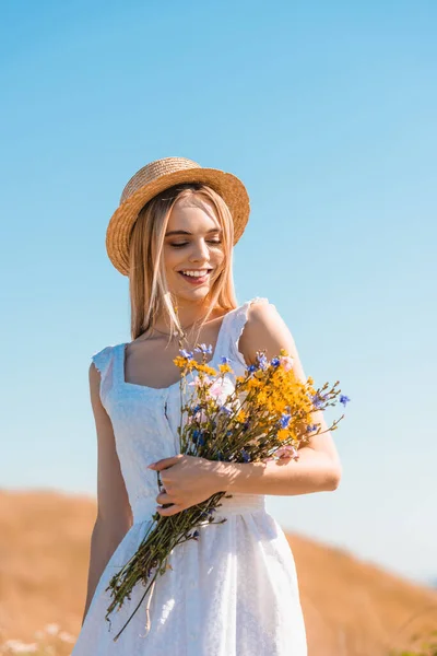 Femme blonde en robe blanche et chapeau de paille tenant bouquet de fleurs sauvages contre le ciel bleu — Photo de stock