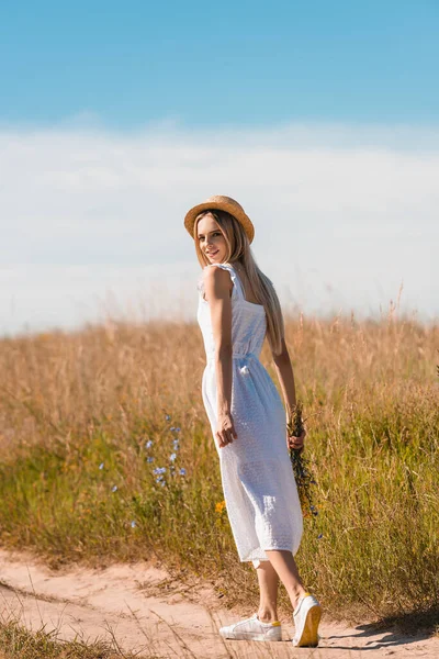 Joven mujer rubia en sombrero de paja y vestido blanco sosteniendo flores silvestres y mirando a la cámara mientras camina por la carretera en el campo - foto de stock