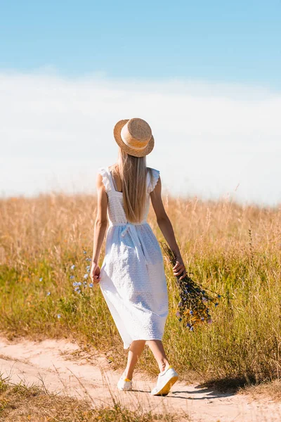 Back view of blonde woman in white dress and straw hat walking on road in field with bouquet of wildflowers — Stock Photo