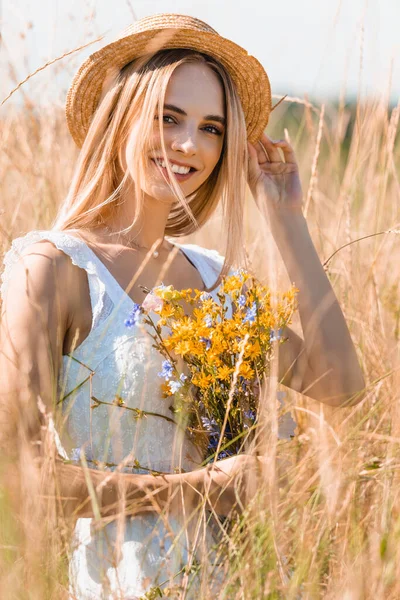 Selective focus of young blonde woman with wildflowers touching straw hat while looking at camera in grassy field — Stock Photo