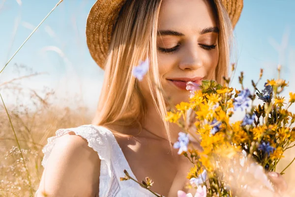 Selective focus of sensual woman in straw hat smelling bouquet of wildflowers with closed eyes — Stock Photo