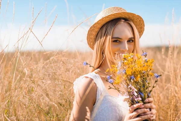 Young blonde woman in straw hat looking at camera while holding wildflowers in grassy meadow — Stock Photo