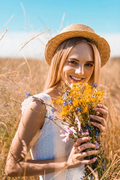 Selective focus of blonde woman in straw hat looking at camera while holding wildflowers in grassy meadow — Stock Photo