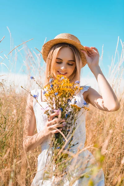 Selective focus of blonde woman in white dress touching straw hat while holding wildflowers in grassy field — Stock Photo