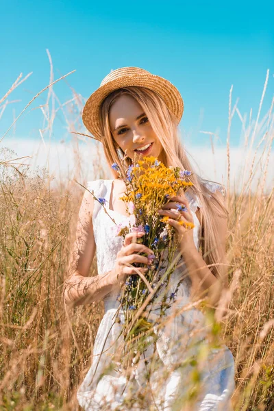 Selektiver Fokus einer sinnlichen Frau in weißem Kleid und Strohhut, die in die Kamera blickt, während sie auf der Wiese Wildblumen hält — Stockfoto