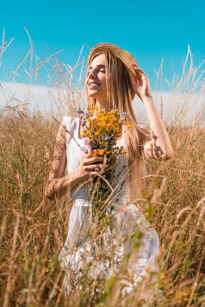 Selektiver Fokus der blonden Frau in weißem Kleid und Strohhut, die einen Strauß Wildblumen hält, während sie Strohhut berührt — Stockfoto