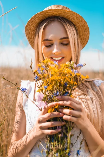 Jovem loira em chapéu de palha segurando buquê de flores silvestres no prado gramado — Fotografia de Stock