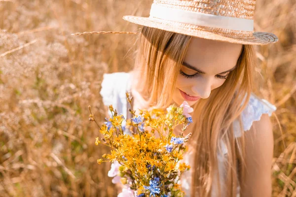 Vue grand angle de femme blonde sensuelle en chapeau de paille tenant bouquet de fleurs sauvages dans prairie herbeuse — Photo de stock