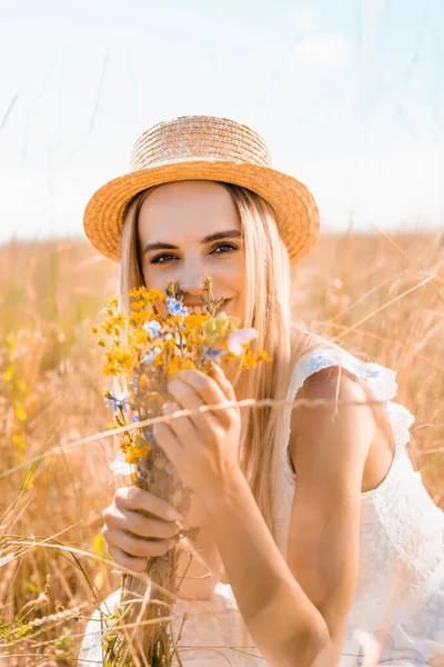 Selective focus of blonde woman in straw hat looking at camera while holding wildflowers in grassy meadow — Stock Photo